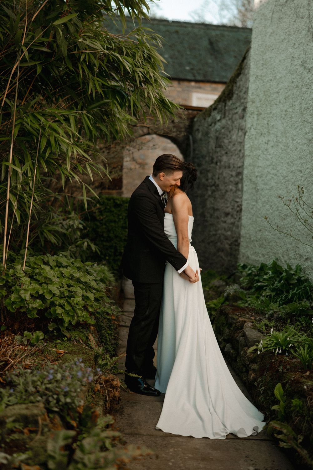 Bride and groom embrace in a lush garden pathway, wearing white gown and black tuxedo at their wedding.