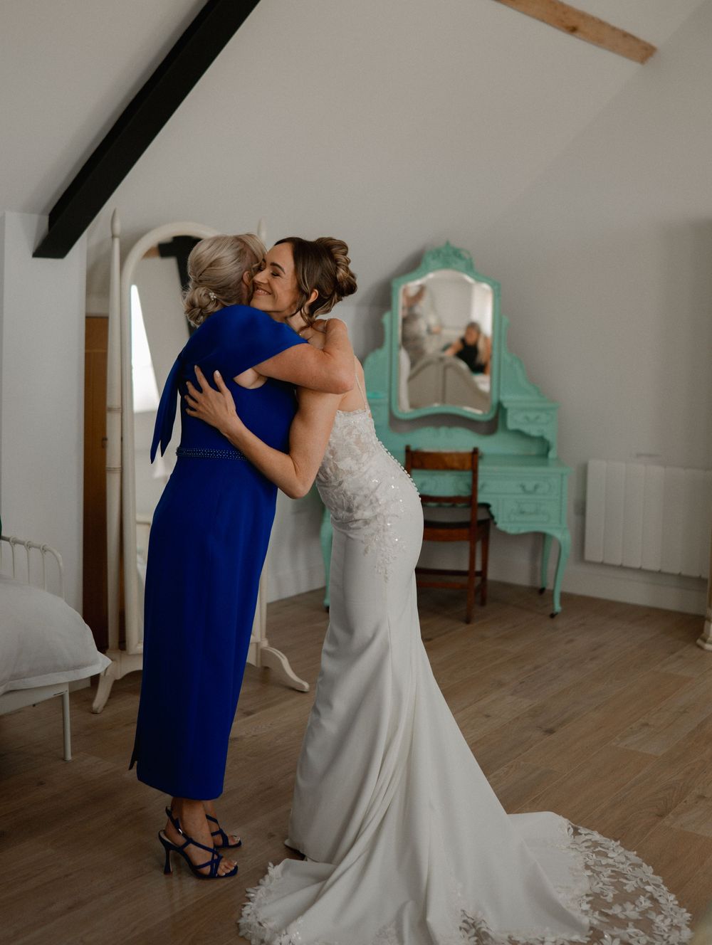 A bride in a white gown embraces a woman in a royal blue dress in a charming room with a mint green vanity mirror.
