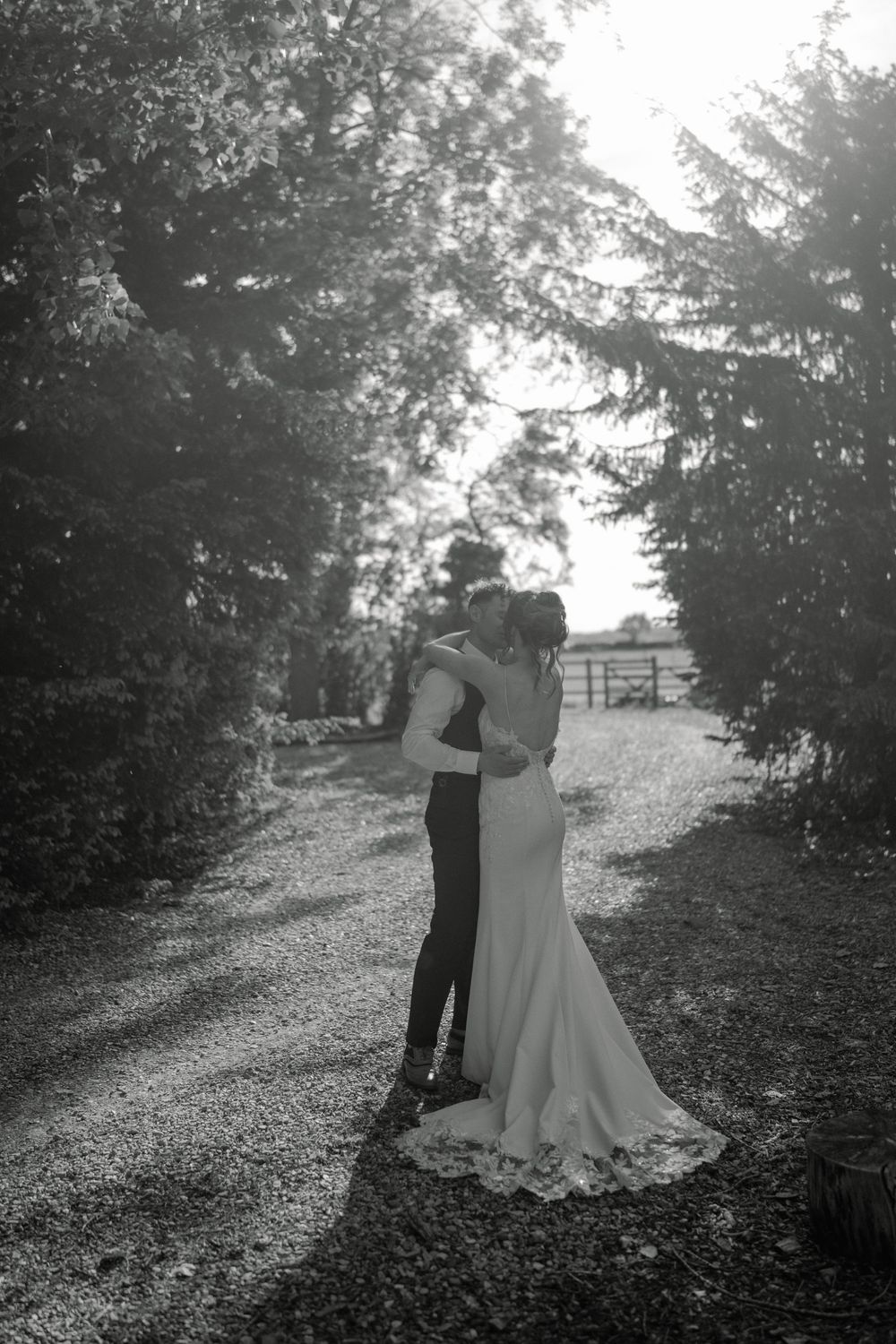 Black and white photo of a bride and groom embracing on a tree-lined gravel path, sharing a romantic moment.
