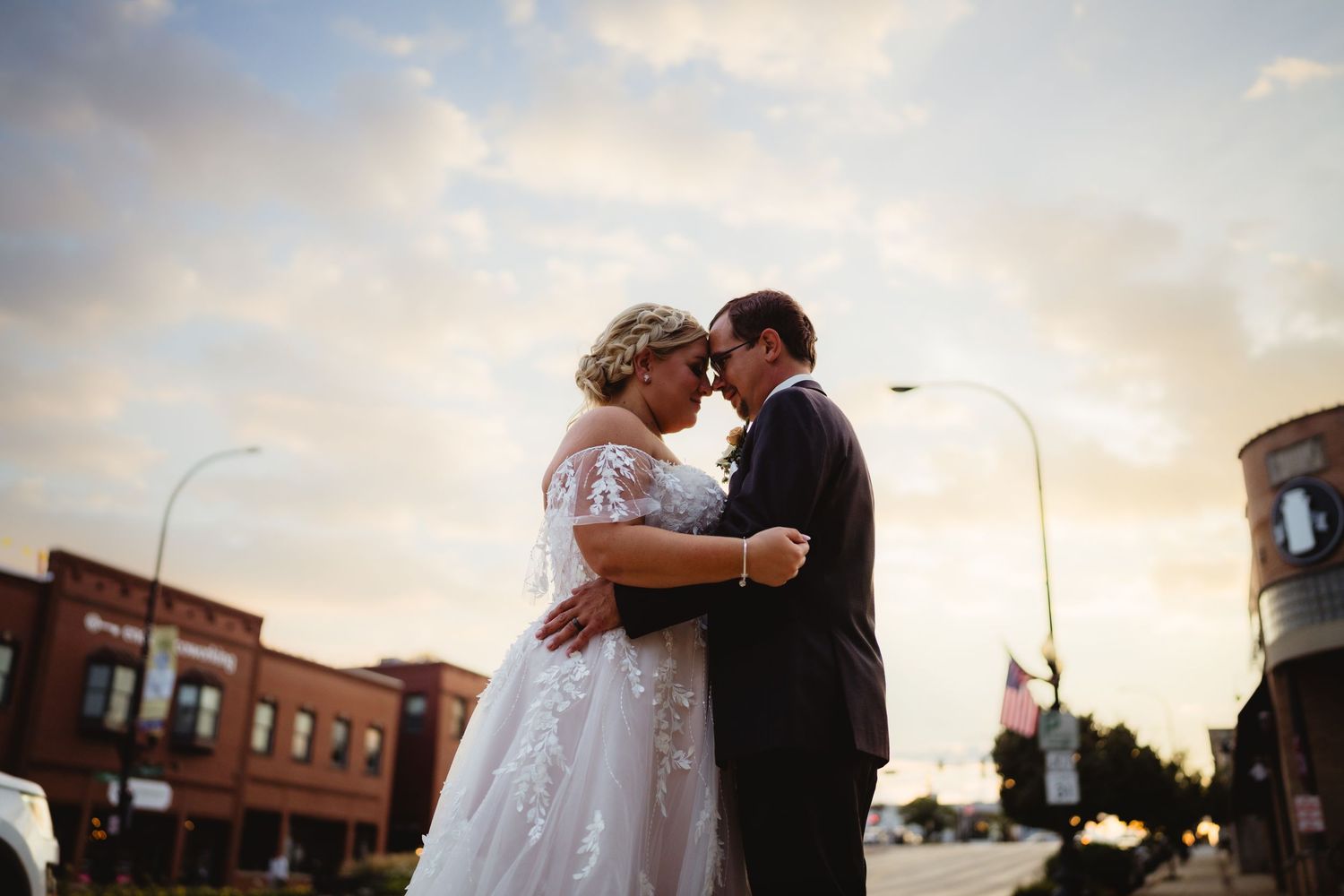 A couple shares a romantic embrace on an urban street corner at sunset with brick buildings in the background.