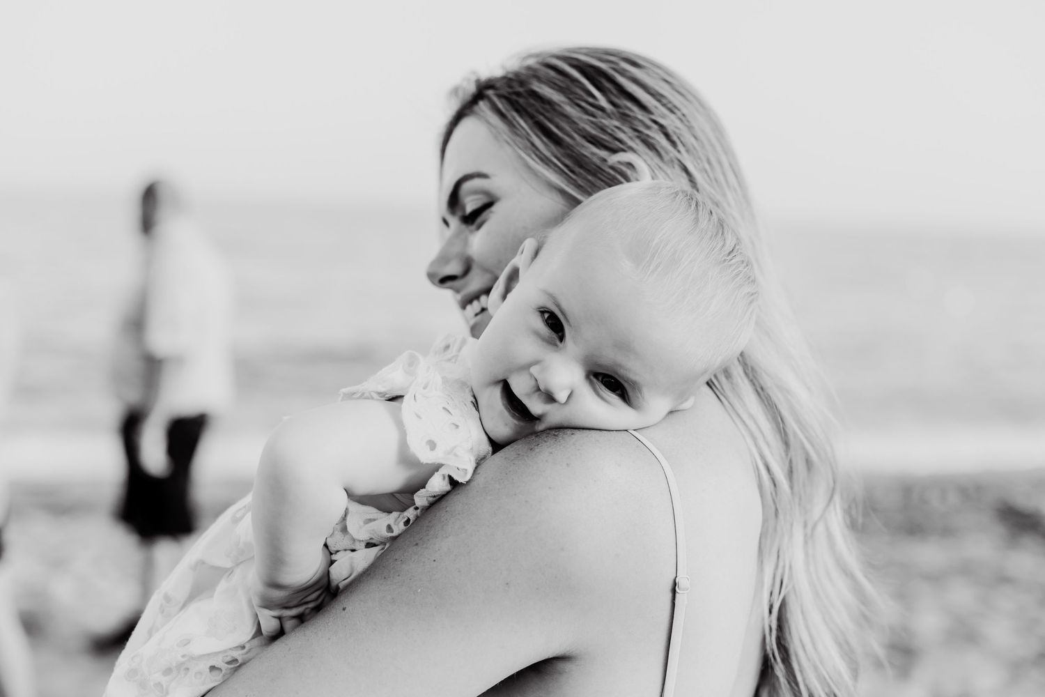 A black-and-white photo of a laughing baby girl on her mother´s shoulders on the beach in Marbella.