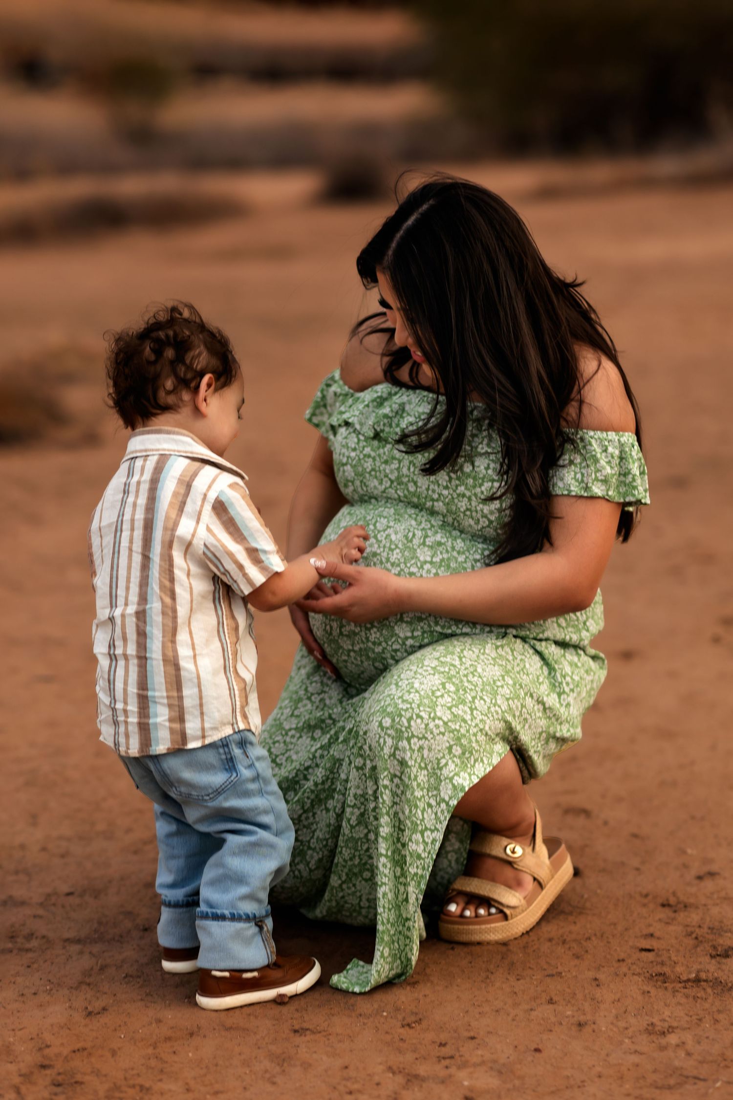 A small child tenderly holds hands with someone wearing a flowing green floral dress while standing on rustic ground.