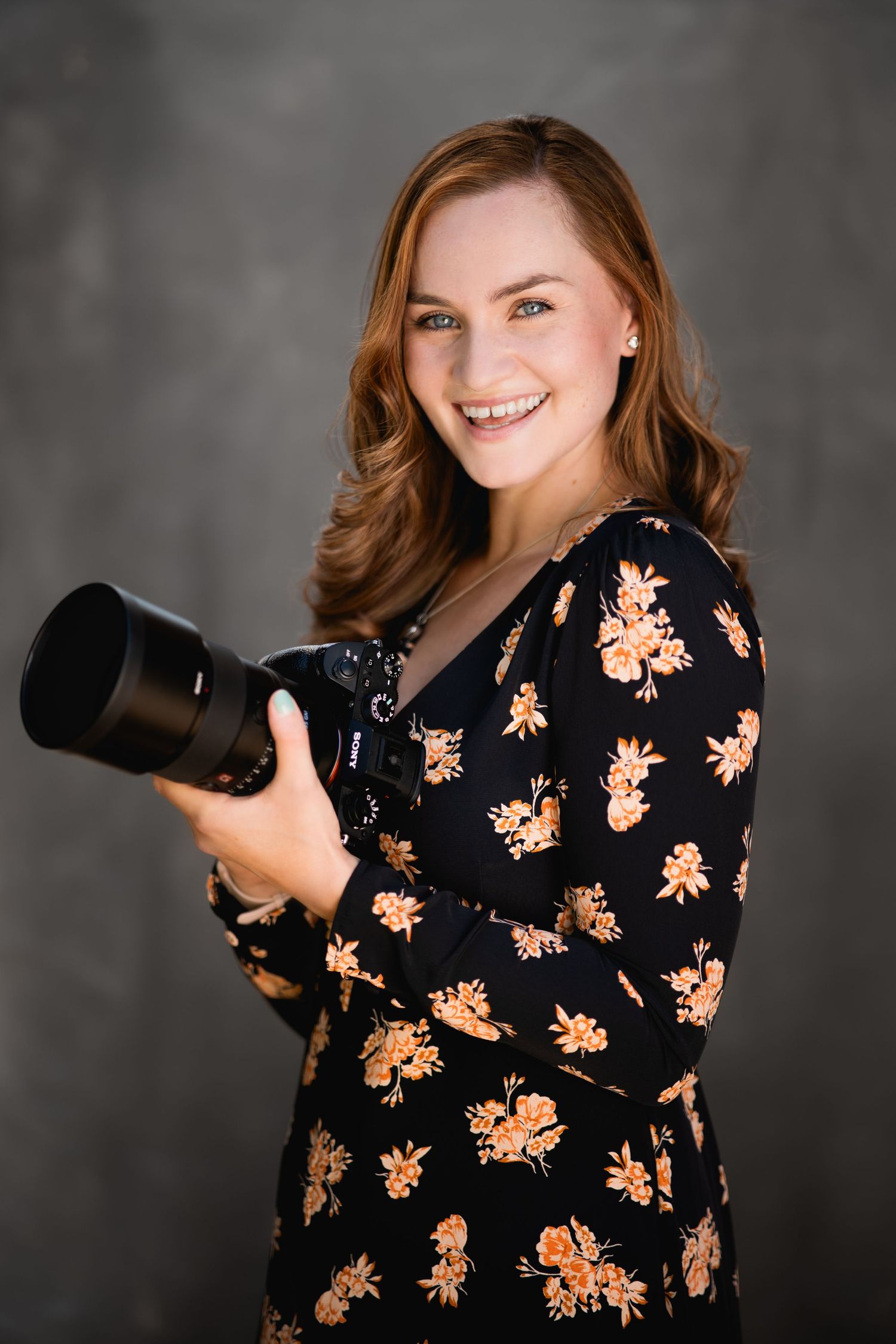 Professional photographer holds a large camera lens while wearing a black floral dress against a dark grey backdrop.