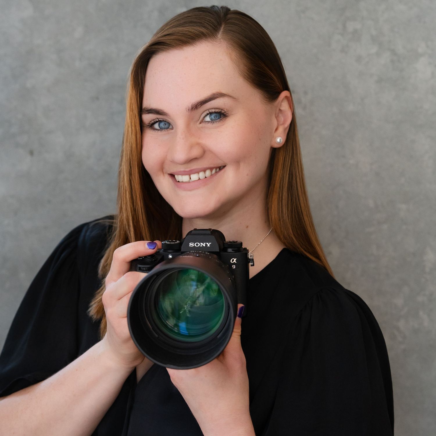 Professional photographer holds a large camera lens while wearing a black floral dress against a dark grey backdrop.
