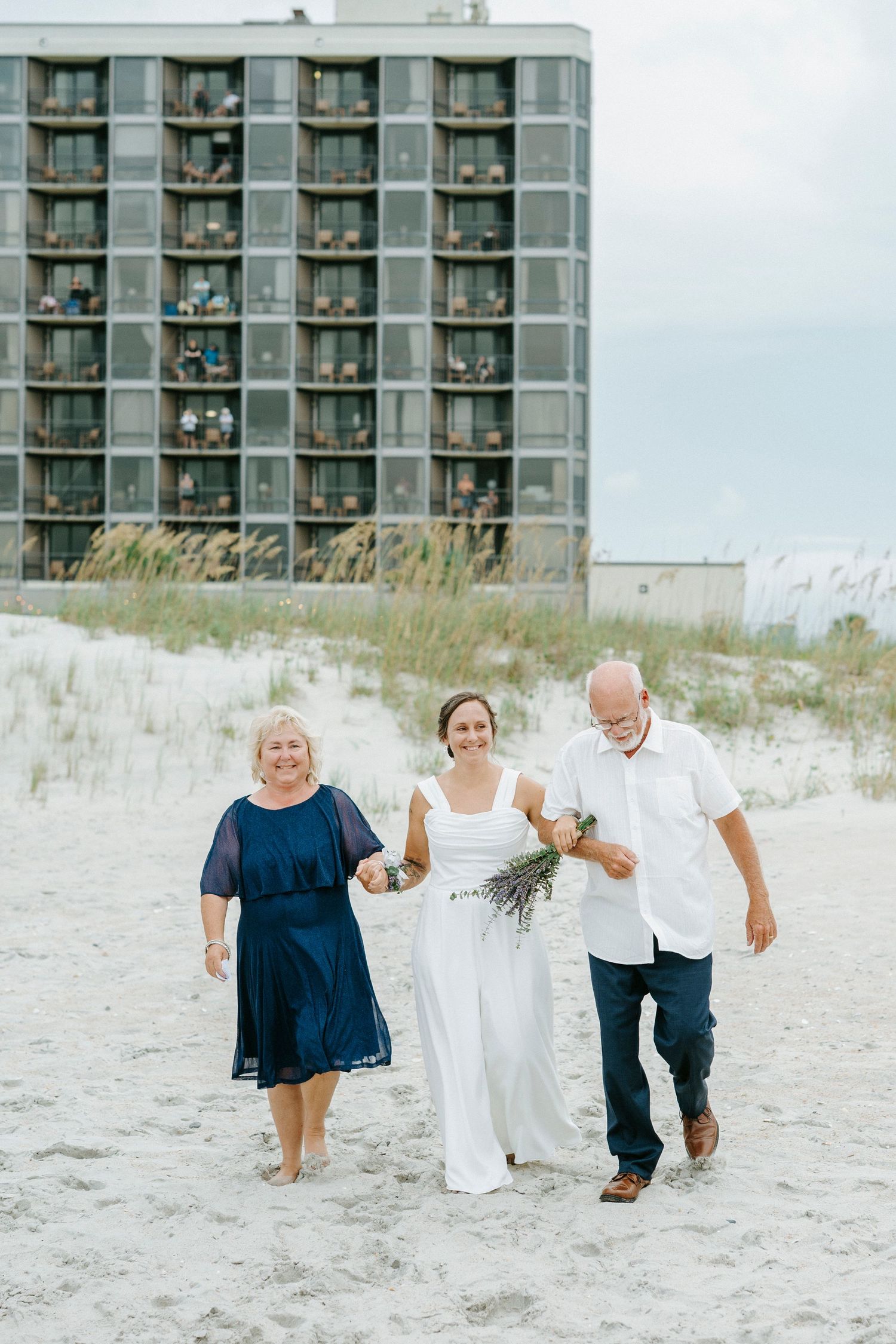 Wedding guests playfully running and dancing across white sandy beach with oceanfront buildings behind them.