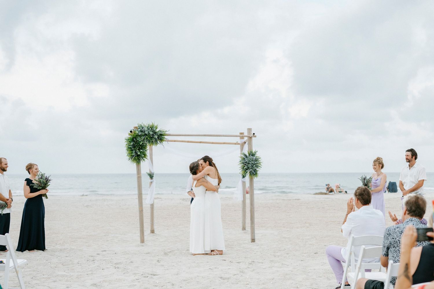 Two people share their first kiss as newlyweds under a wooden arch decorated with greenery on a cloudy beach.
