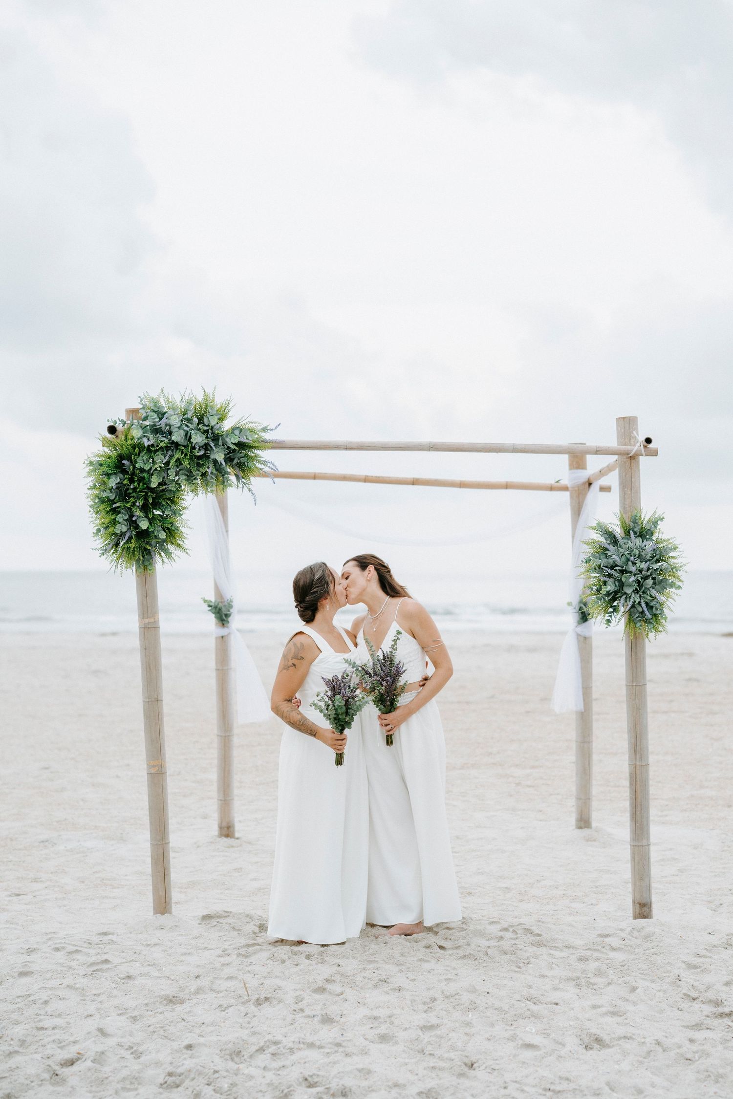 Minimalist beach wedding ceremony setup with wooden posts and greenery decorations against ocean backdrop.