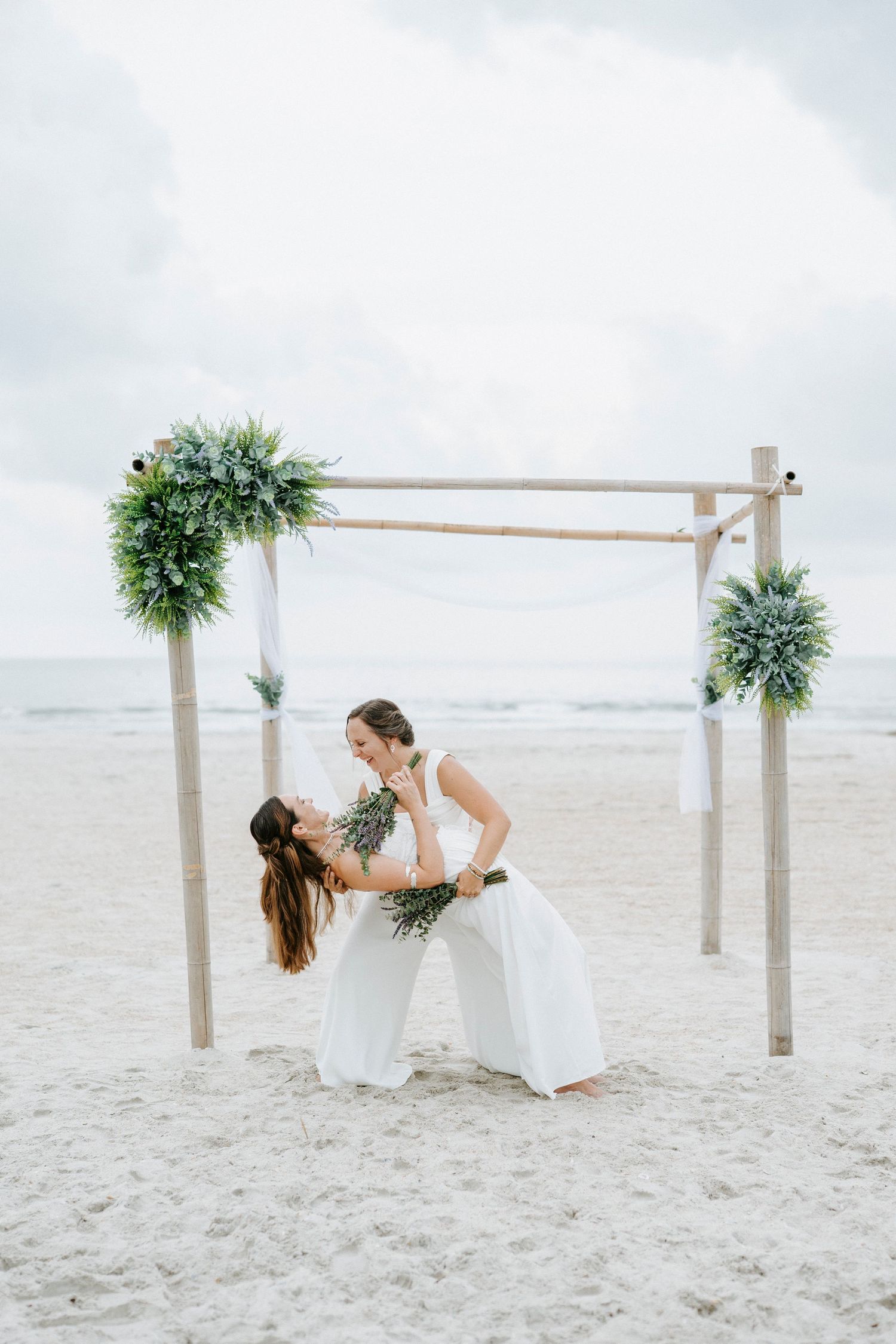Two people in white dresses embrace on a beach under a wooden wedding arch with greenery.