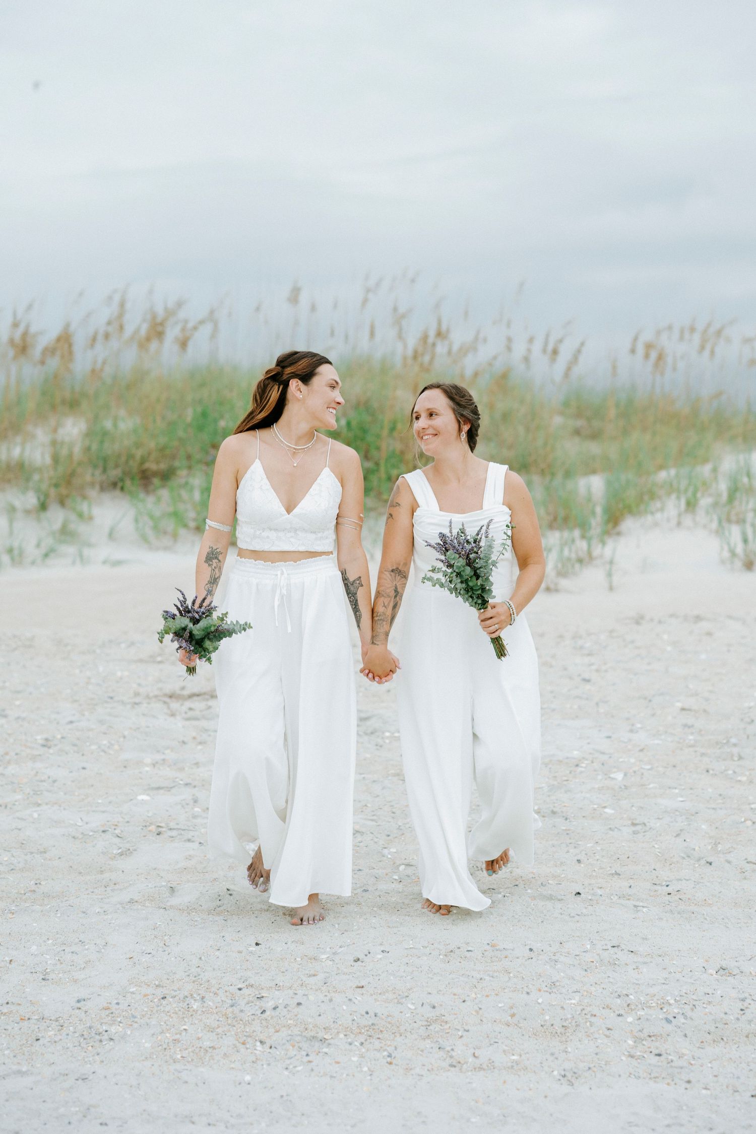 Two people in white dresses hold hands while walking on a sandy beach with sea grass in the background.
