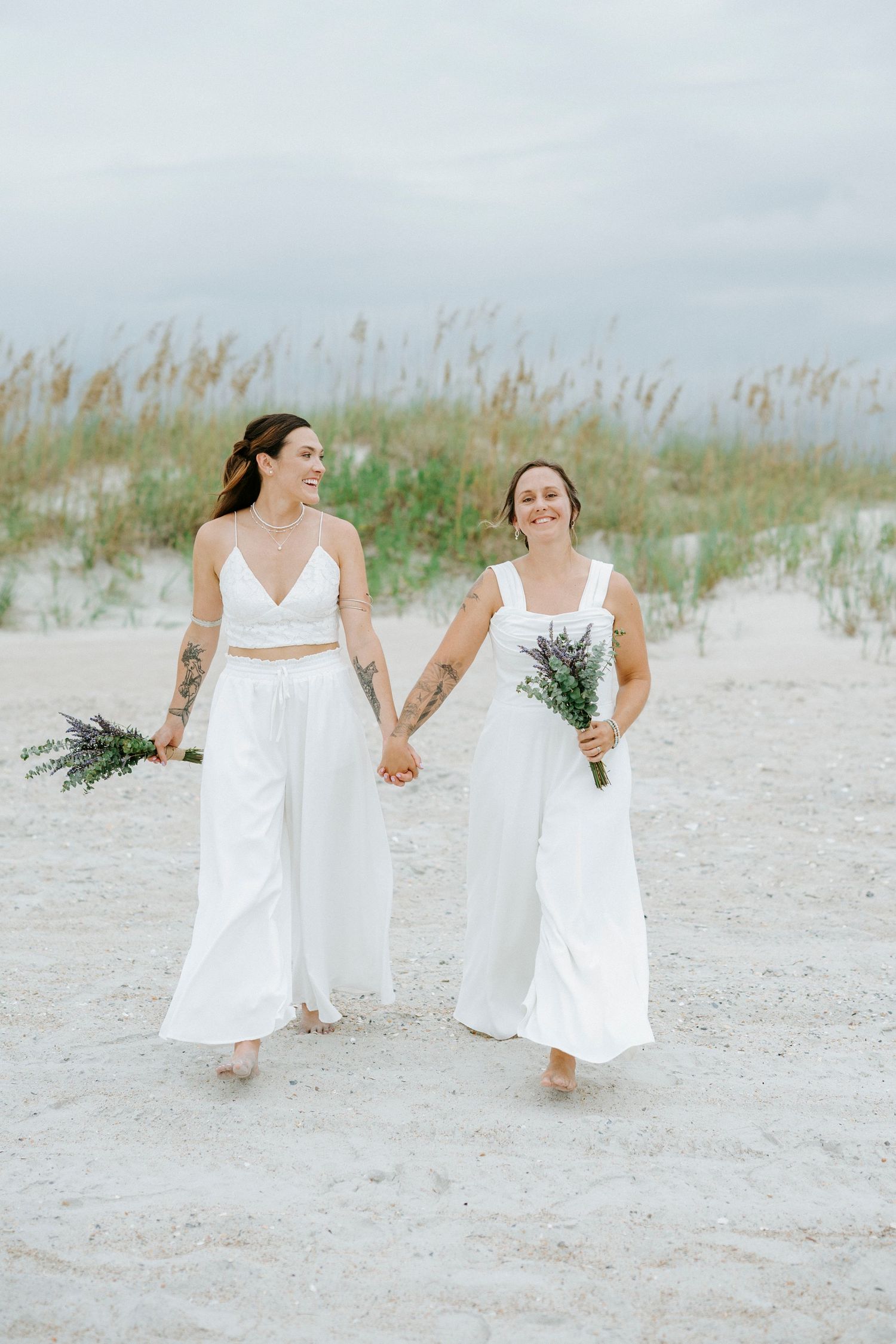 Couple in white dresses holding hands while walking along a peaceful sandy beach with sea oats in the background.