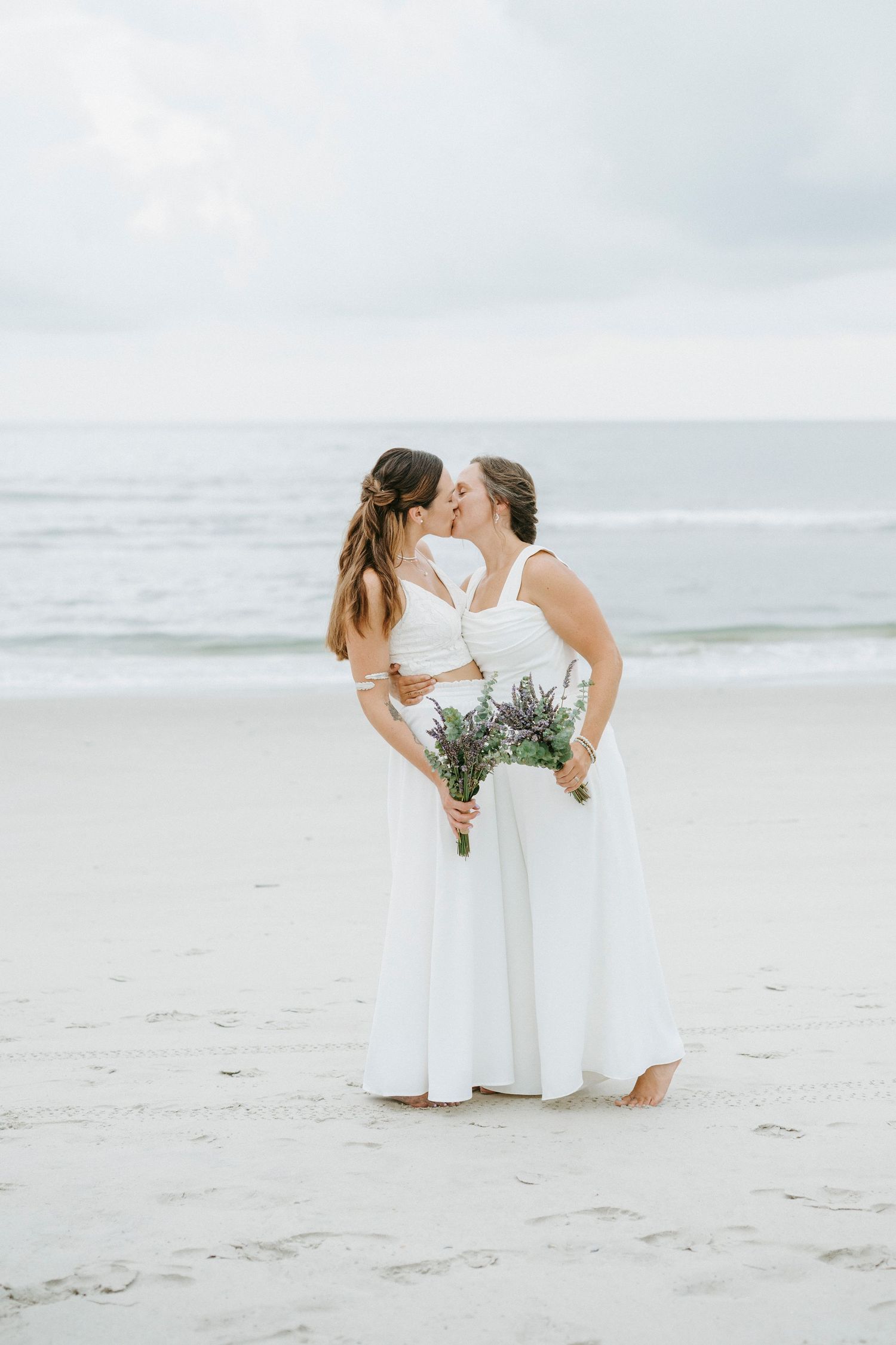 A romantic beach wedding photo shoot showing two people in elegant white dresses sharing an intimate moment by the ocean.