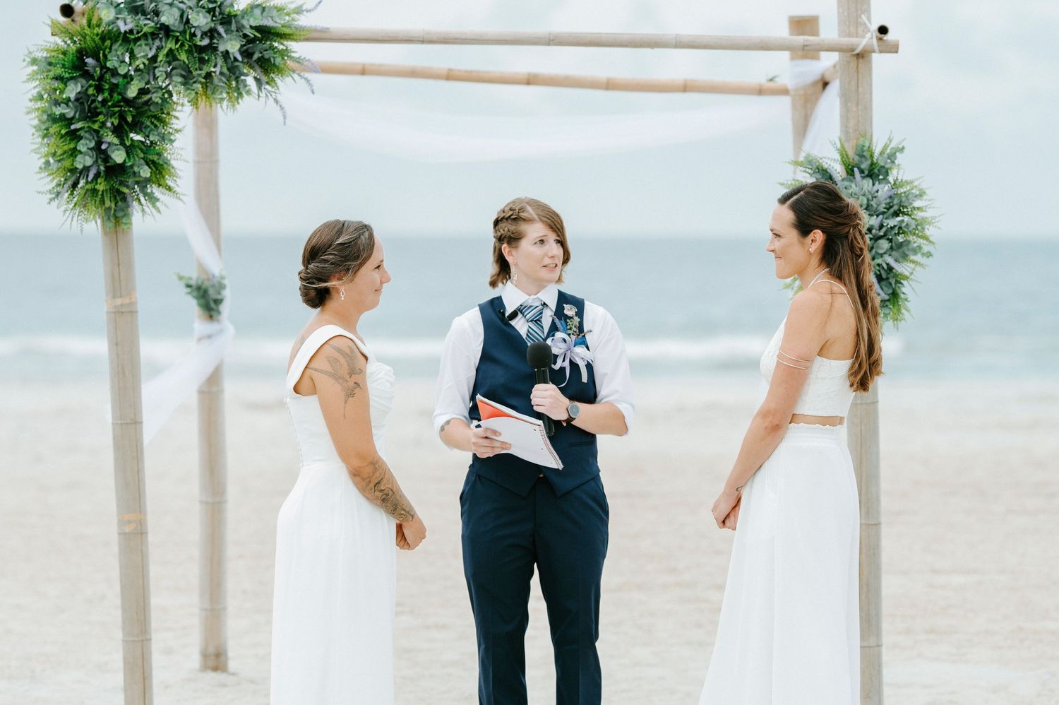 Three people stand under a bamboo wedding arch on a beach during an intimate ceremony.
