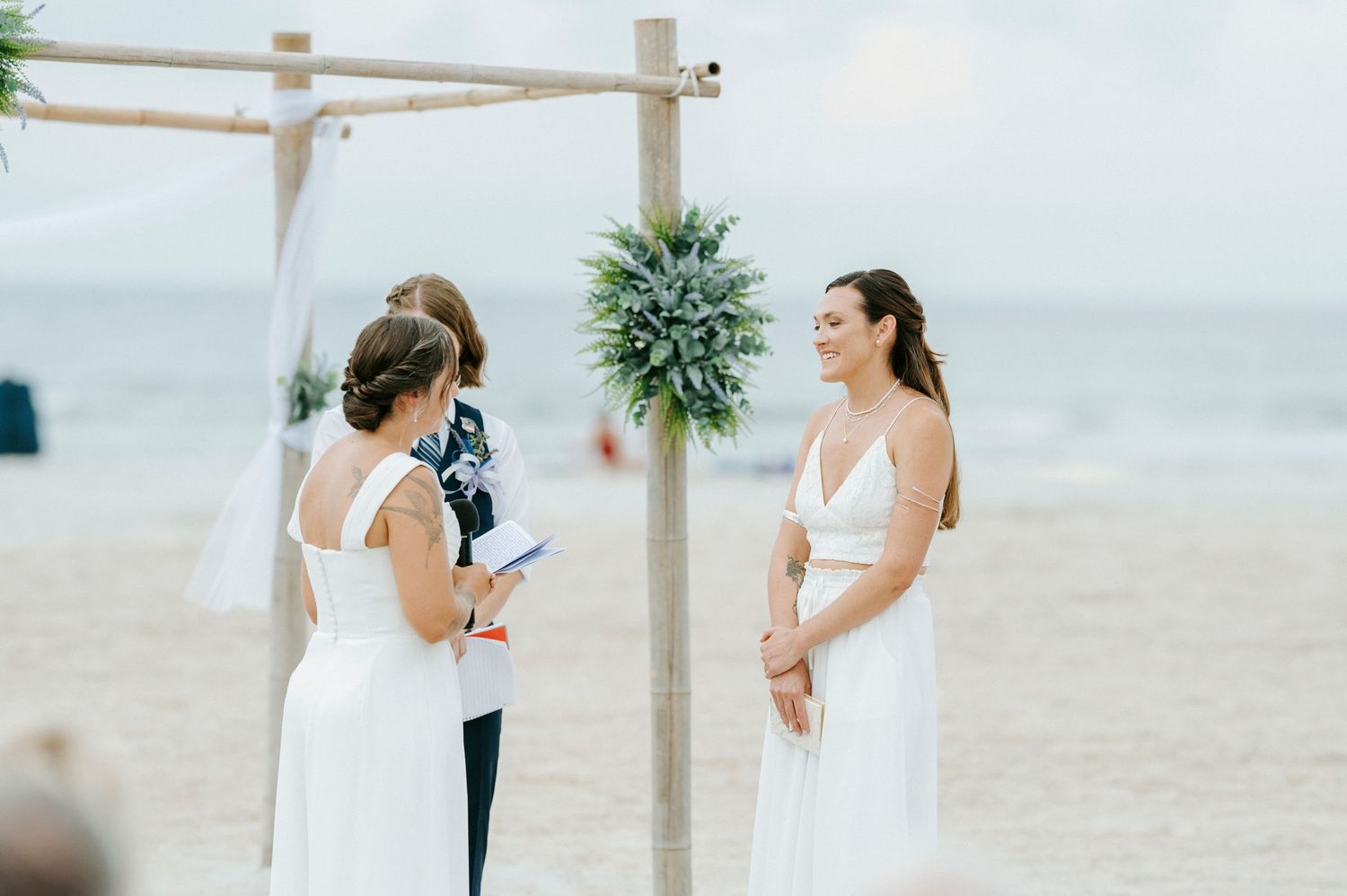 Two people embrace during a beach wedding ceremony under a wood arbor decorated with white fabric and greenery.