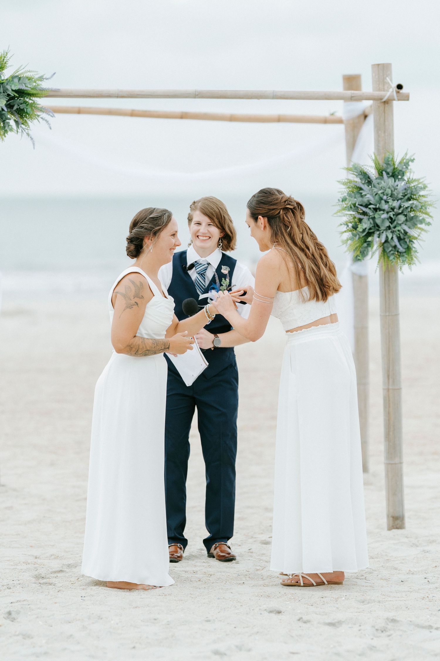 Wedding party gathers around a rustic wooden arch on a serene beach.