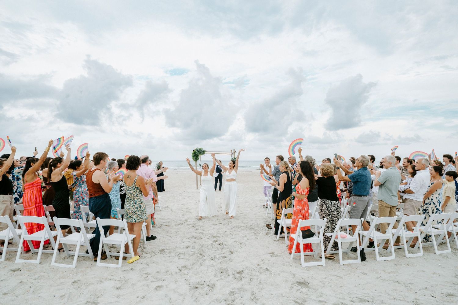 Large group of wedding guests celebrating on beach with white chairs and cloudy sky backdrop.