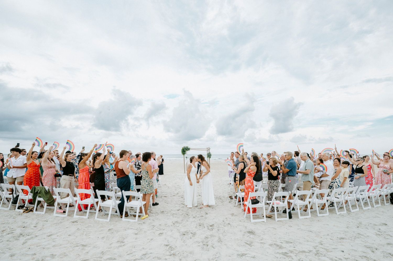 Wedding guests seated in white chairs on a sandy beach for an oceanfront ceremony.