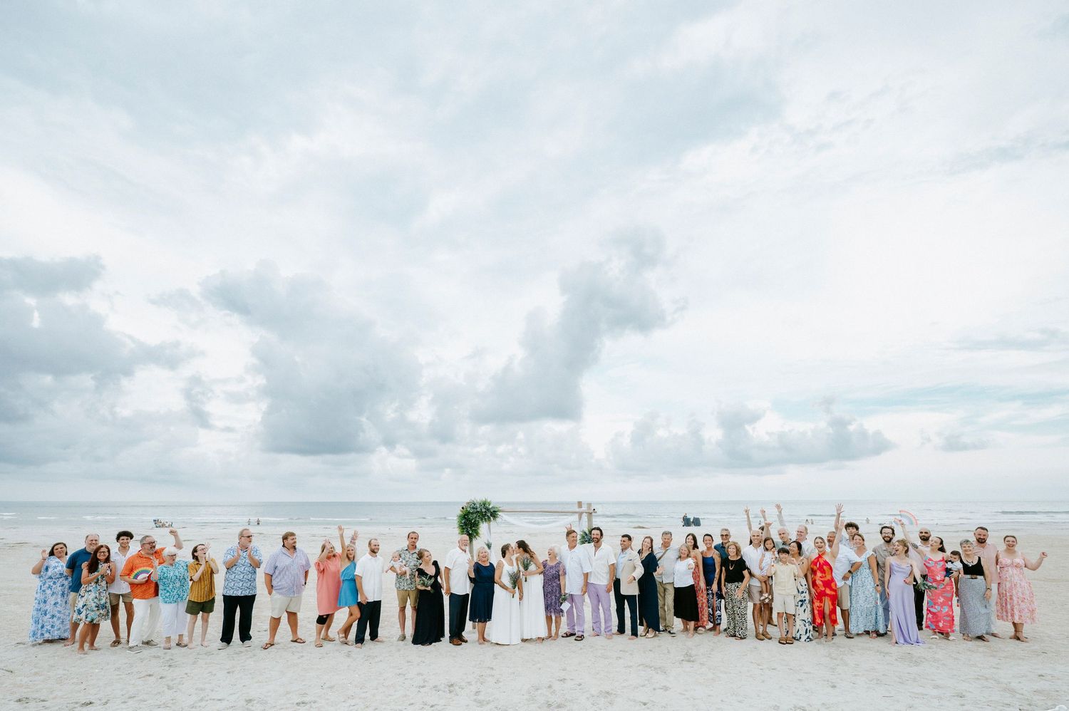 Large group wedding photo with guests lined up on beach under cloudy sky.