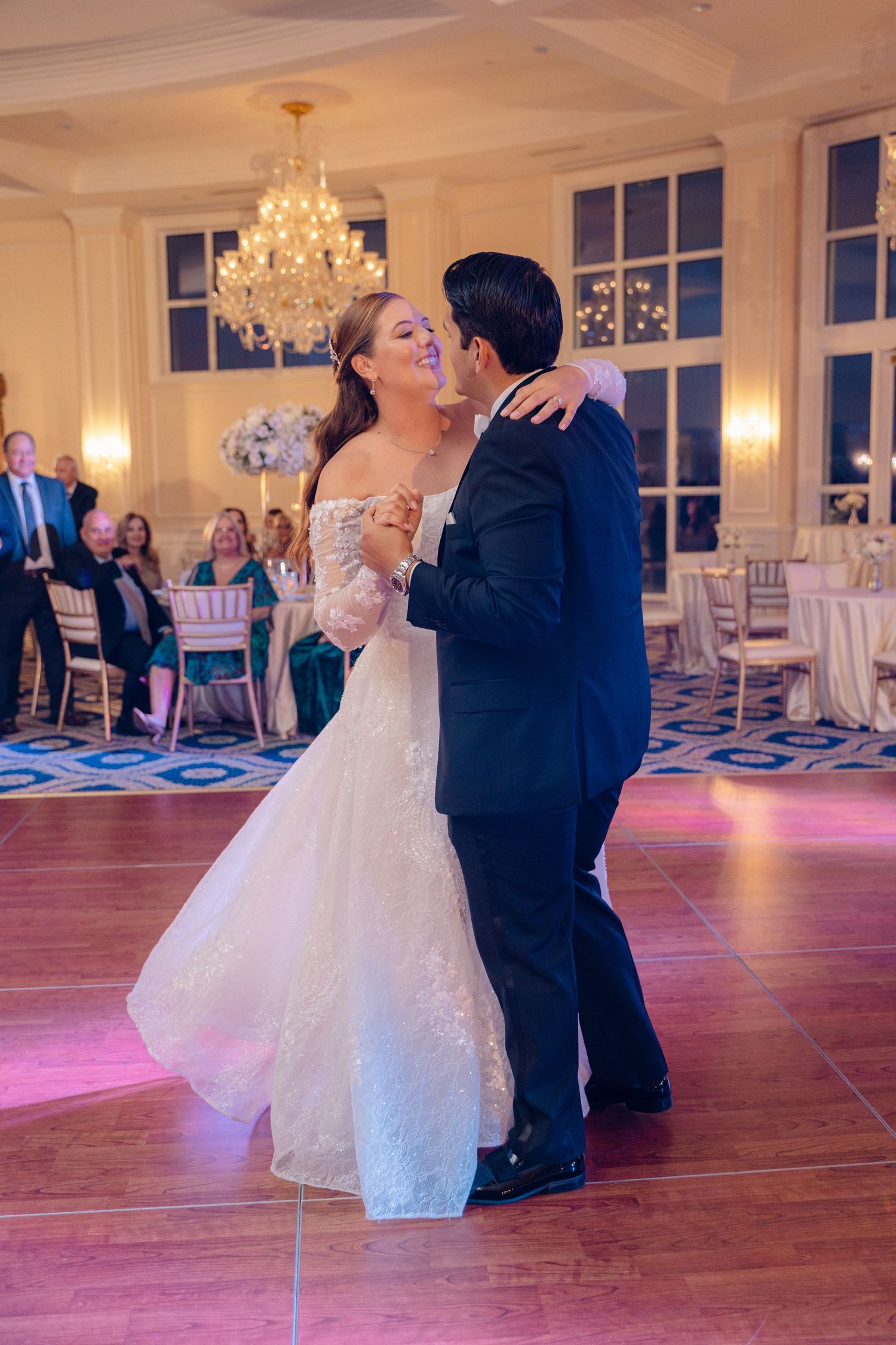 Couple shares their first dance in an elegant ballroom with crystal chandeliers and romantic pink lighting.