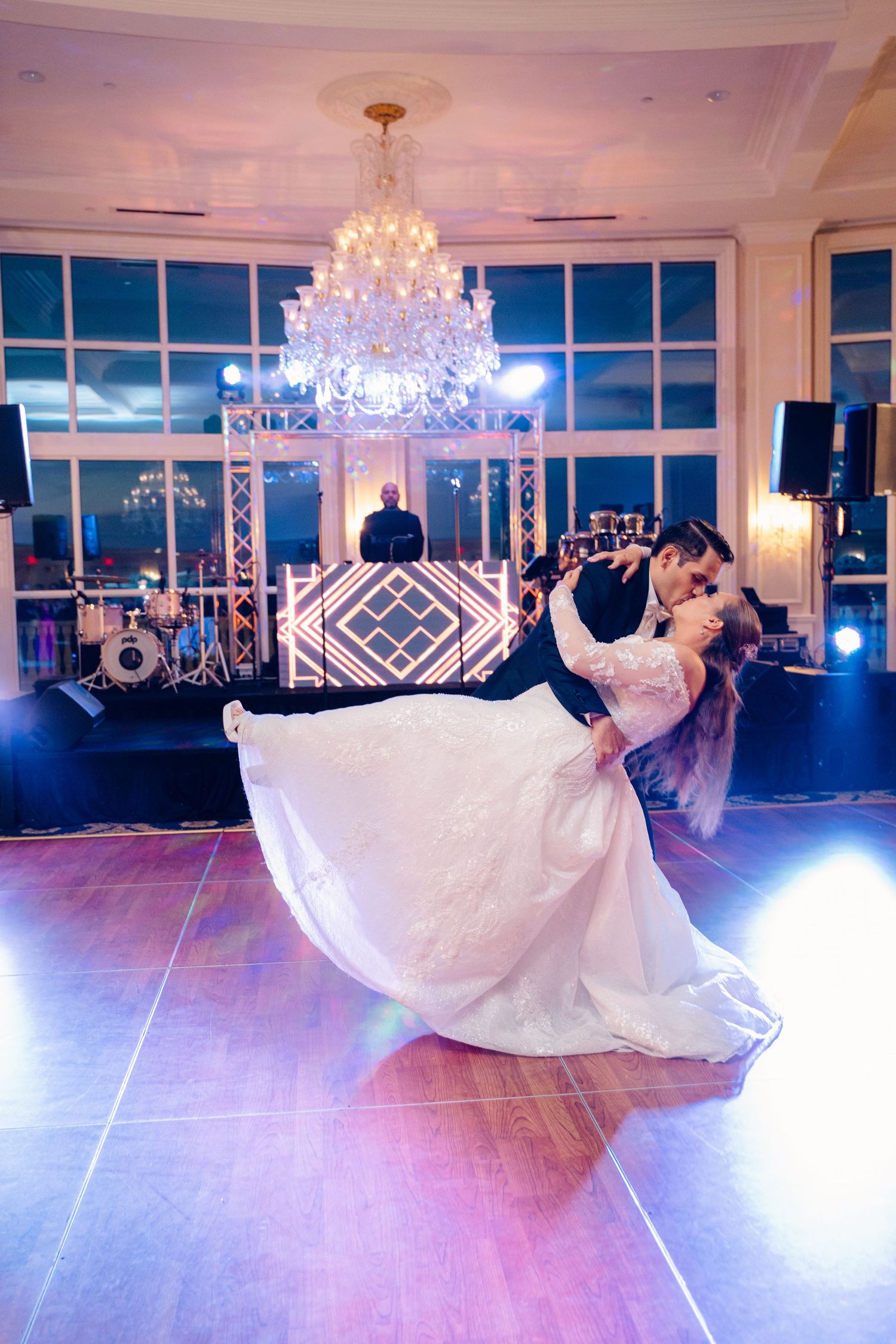 A couple shares a romantic dance dip under a crystal chandelier in an elegant ballroom with blue lighting and windows.