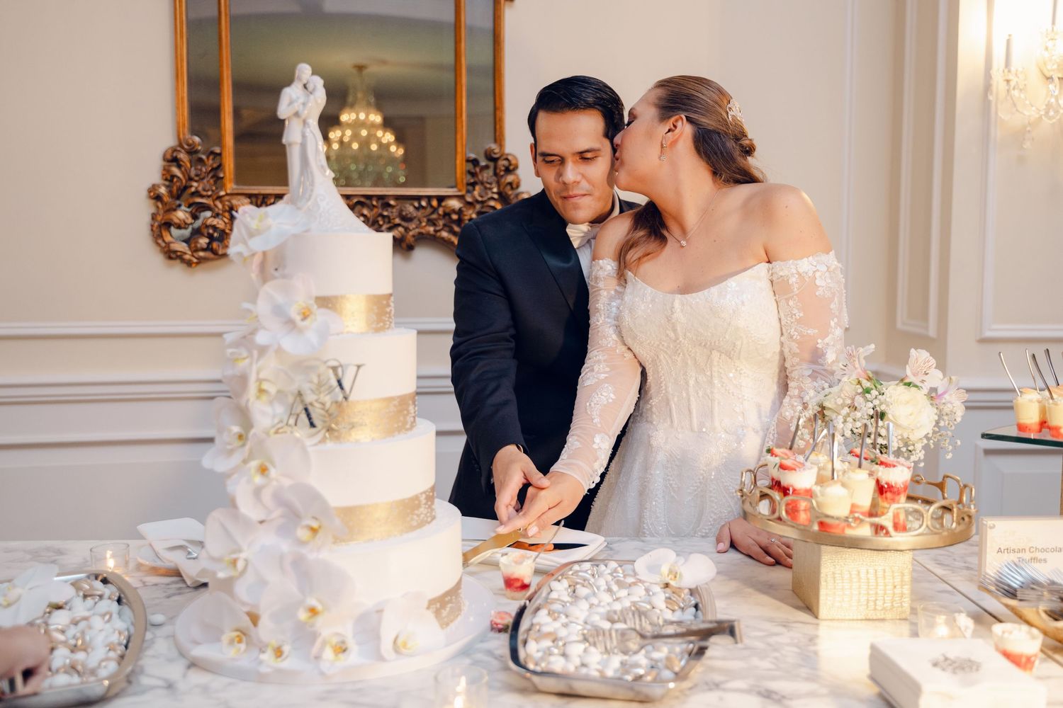 A wedding couple cuts into their elegant white tiered cake decorated with delicate floral details at an ornate reception venue.
