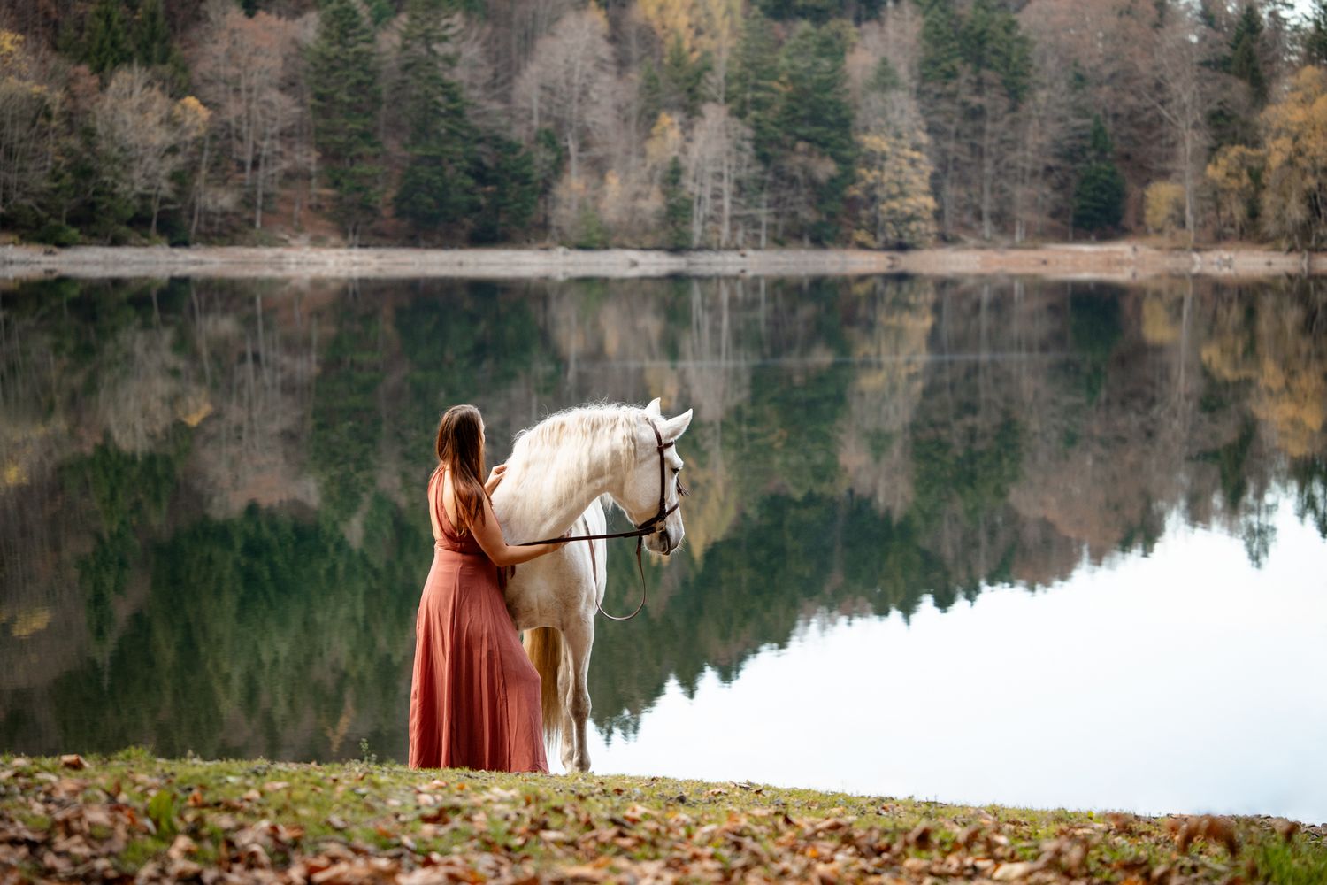 A woman in a red dress stands beside a white horse near a calm reflective lake in autumn.