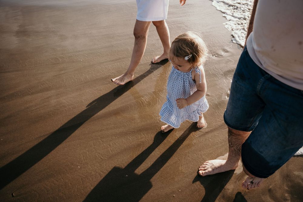 Toddler girl in striped dress walking barefoot on sandy beach between parents casting long shadows.