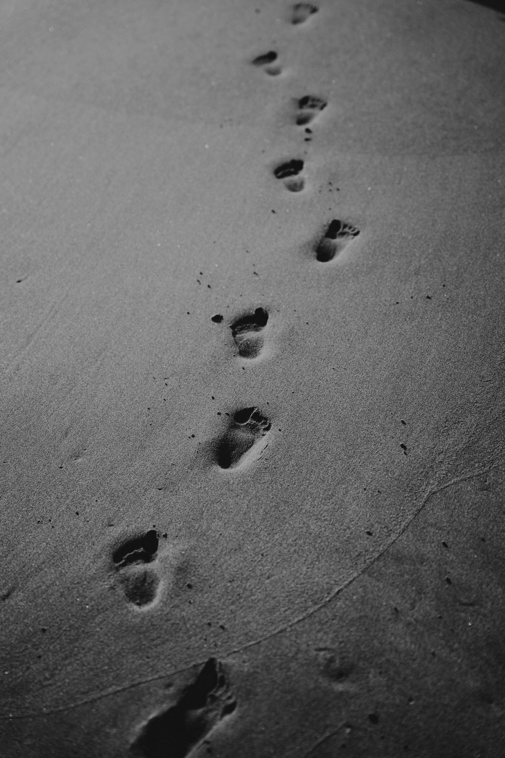 Black and white artistic photo of barefoot footprints trailing across smooth sandy beach.