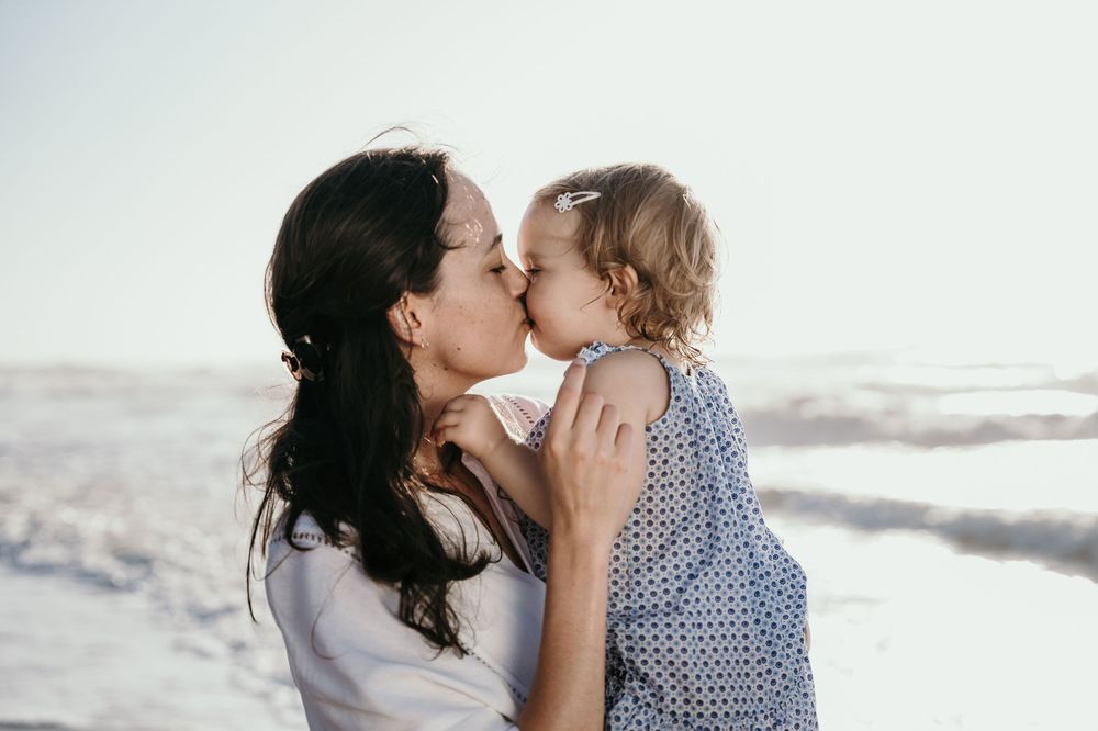 A mother tenderly kisses her young toddler daughter on a bright sunny beach day.