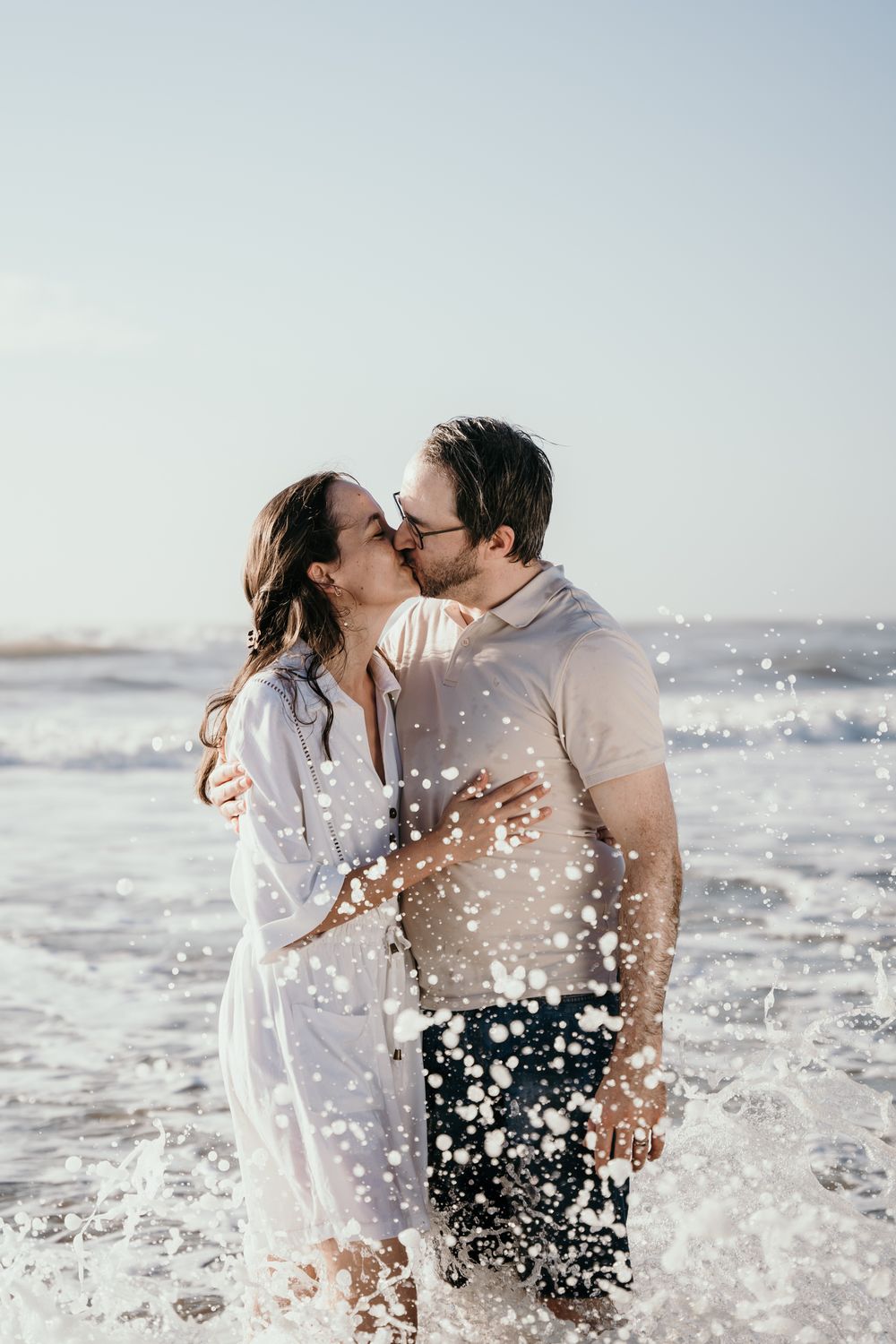 Couple kissing passionately in ocean waves on sunny beach, water splashing around them dramatically.