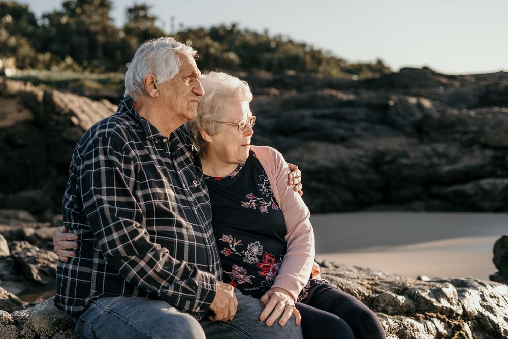 Elderly couple sitting on rocky beach, holding hands and gazing peacefully at the ocean together.