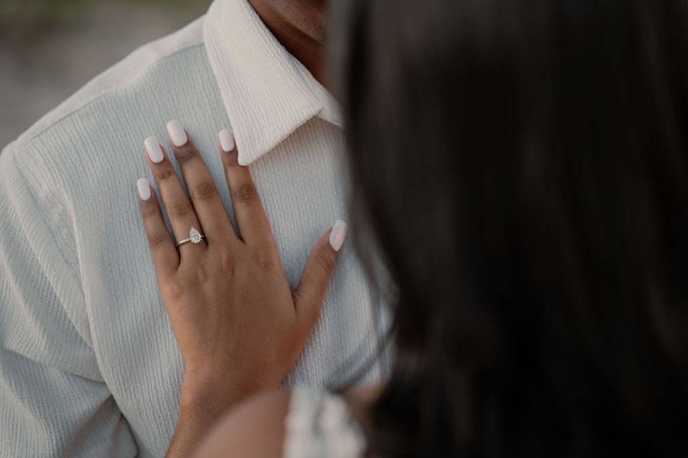 Close-up of womans hand with diamond engagement ring resting on mans white striped shirt.