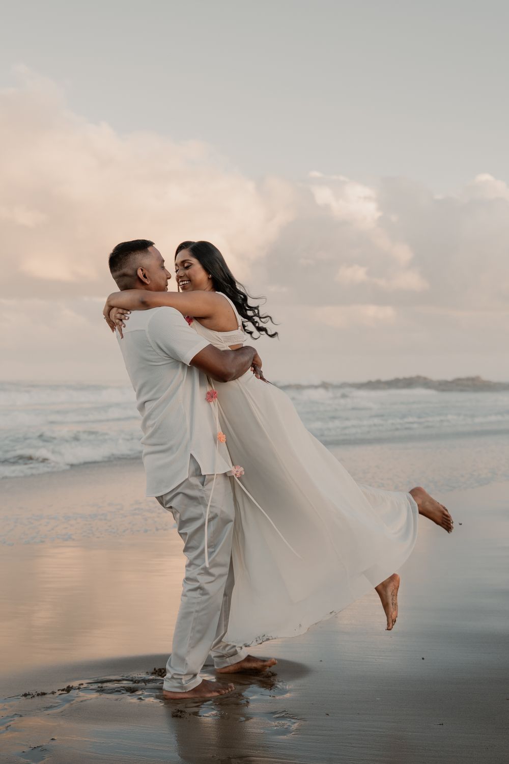 Couple sharing romantic moment on beach at sunset, man lifting woman in white flowing dress near ocean waves.