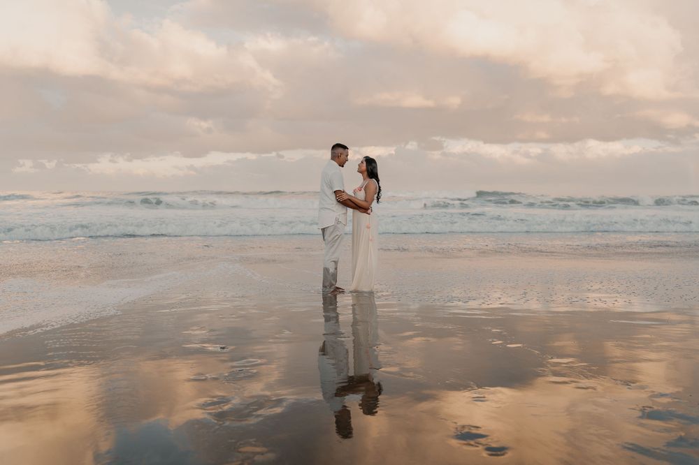 Couple in white outfits standing together on reflective wet beach sand during golden sunset by the ocean.