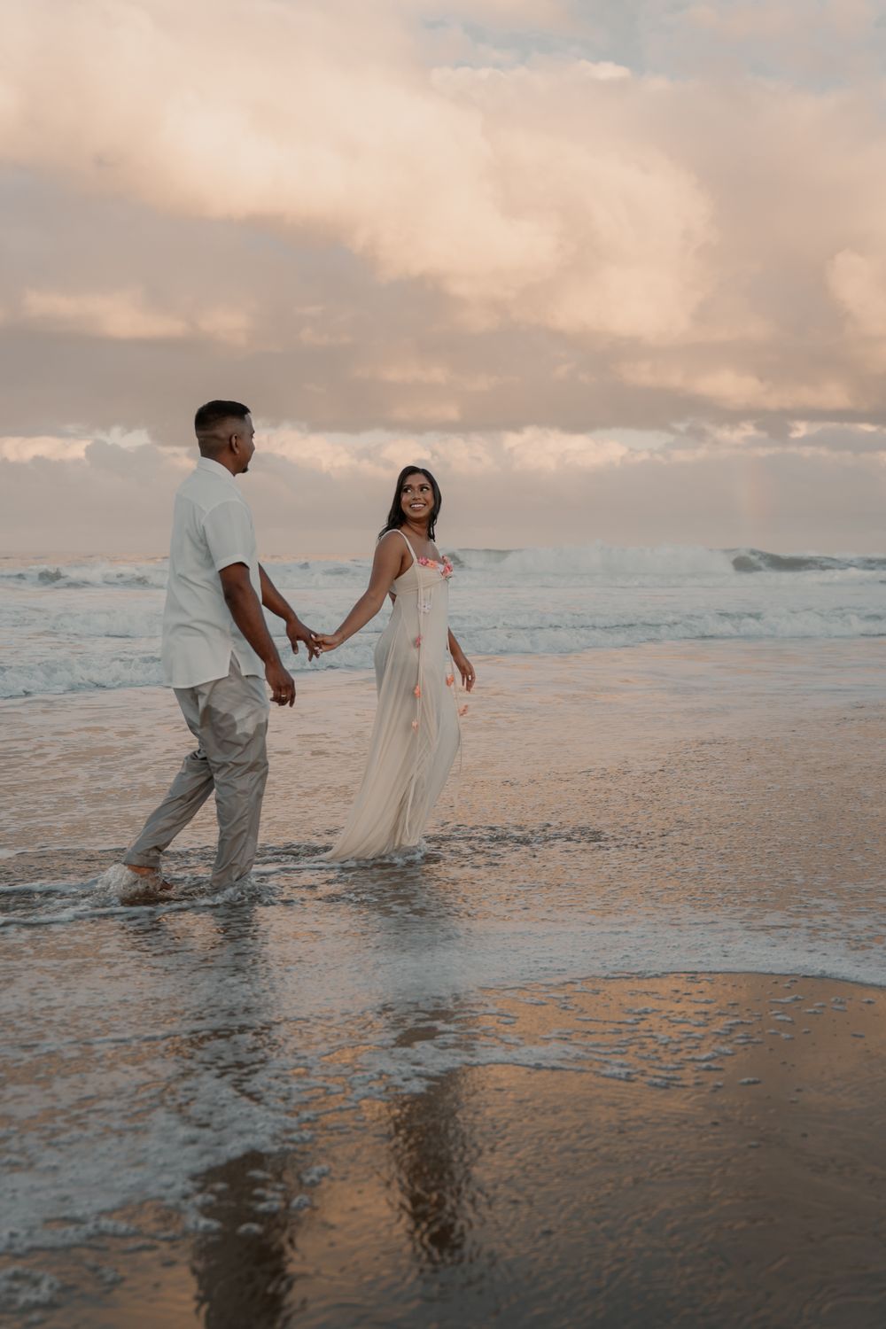 Happy couple holding hands walking barefoot along shoreline at sunset, woman in flowing white dress smiling.