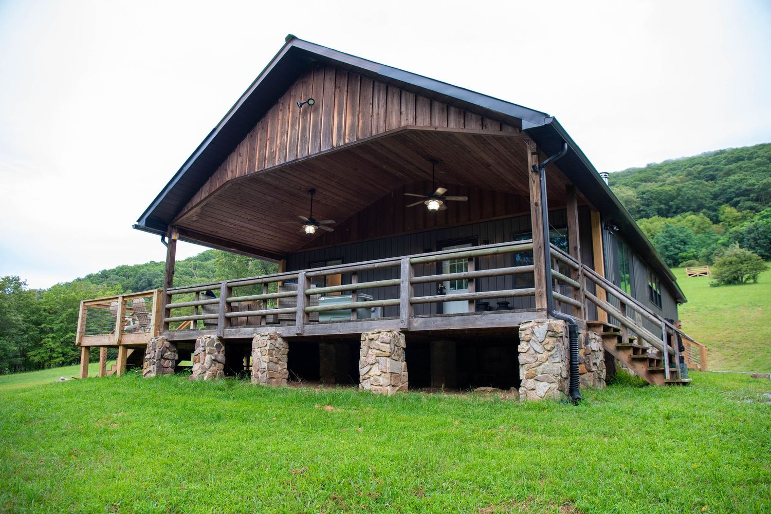 A rustic wooden barn with stone foundation sits nestled on a green hillside in the countryside.