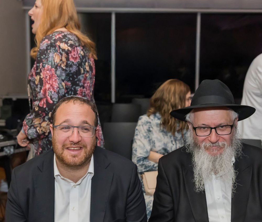 A group of Orthodox Jewish men in traditional black suits and beards smile while gathered at a social event.