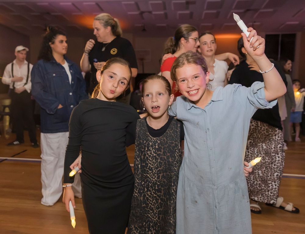 Three young friends pose together at a dance or social event in a wood-floored venue with warm lighting.