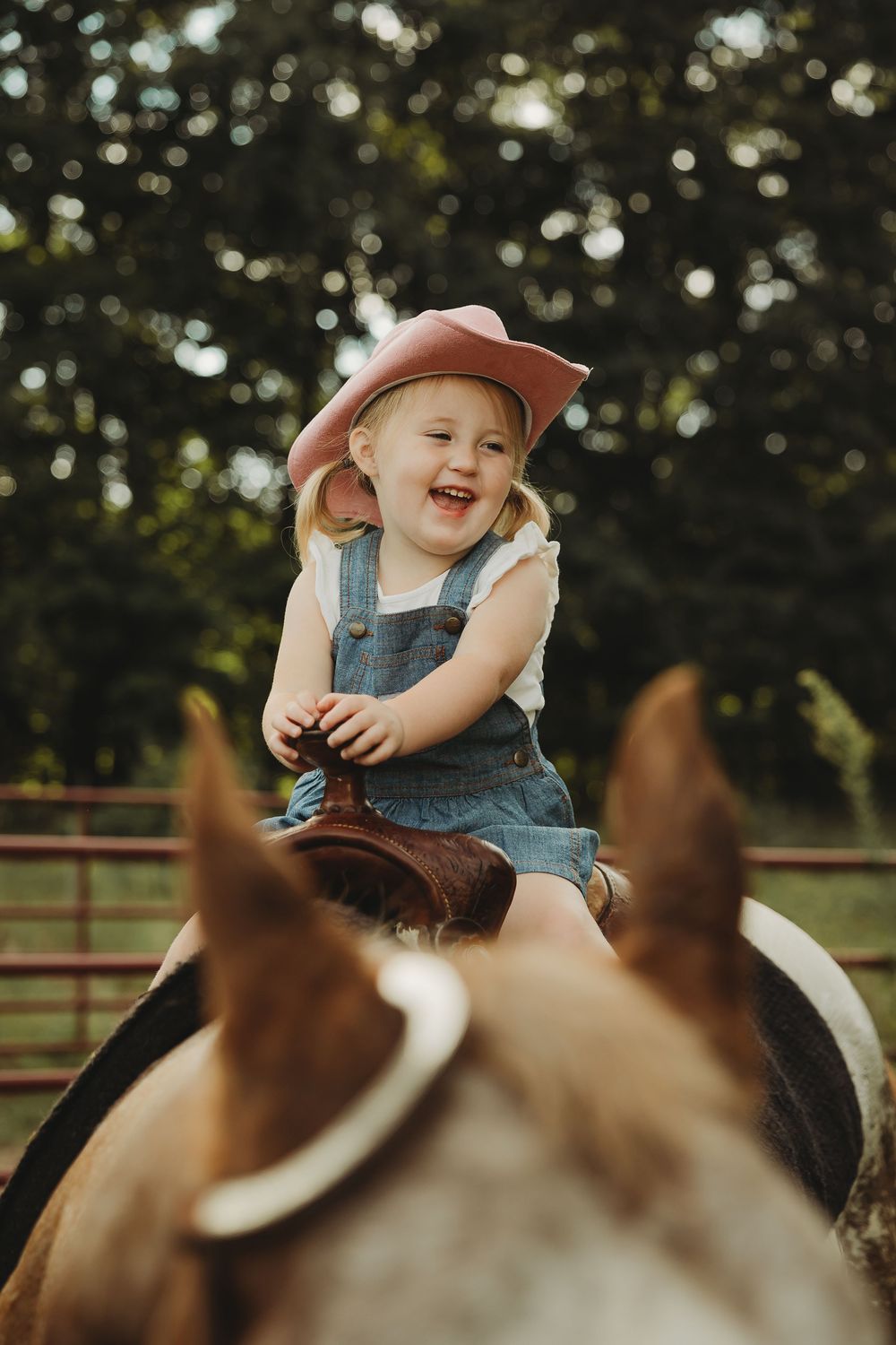 A child wearing denim overalls and a brown hat smiles while riding a horse outdoors.