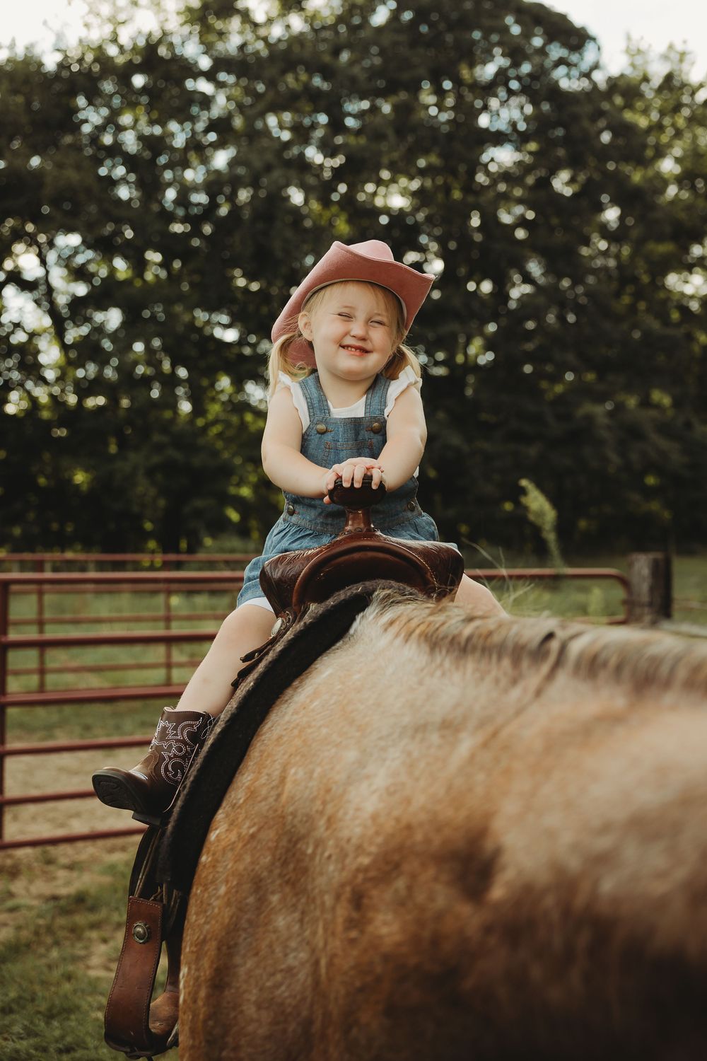 A young child wearing denim overalls and a brown cap sits atop a brown horse in a fenced pasture at sunset.