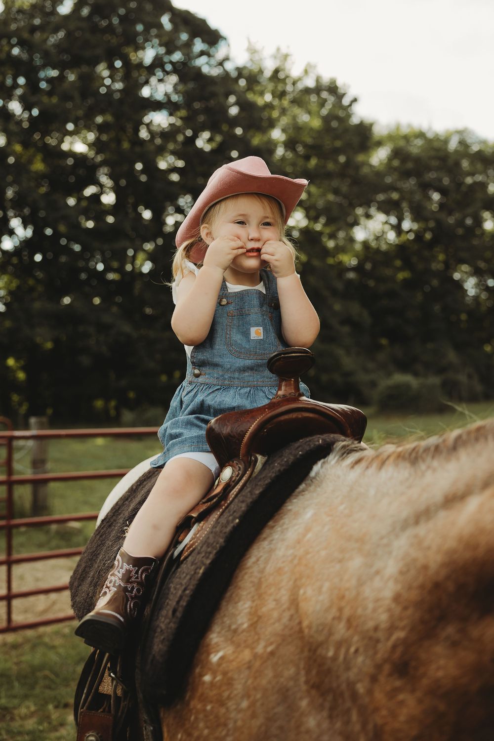 A young child in overalls and hat sits on horseback in a rustic outdoor setting during golden hour.