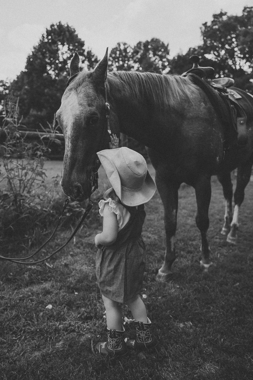 Black and white photo of a person in a dress and hat tending to horses in a rural field setting.