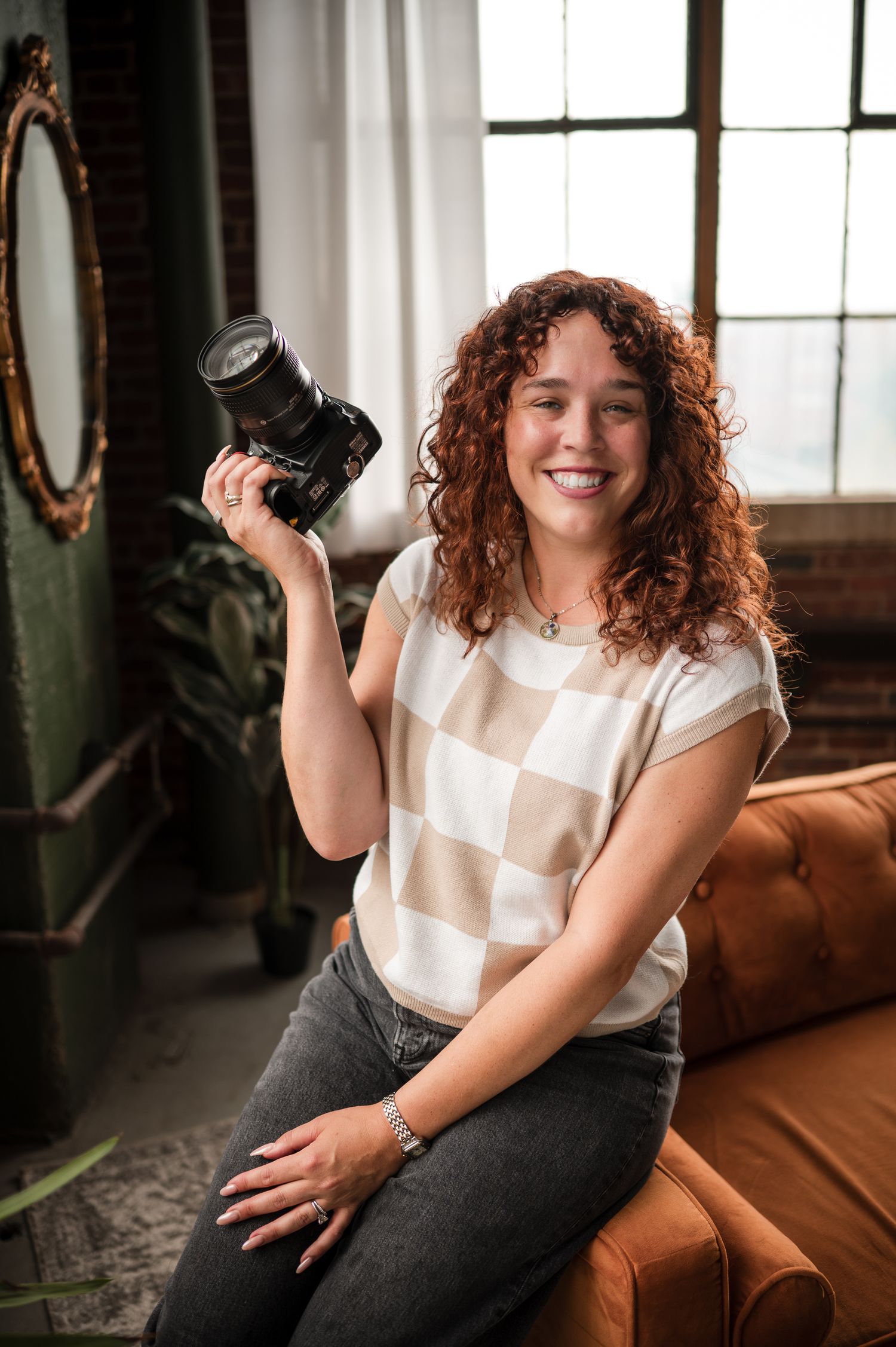 Curly red-haired photographer smiling while holding a camera seated on a brown leather sofa.