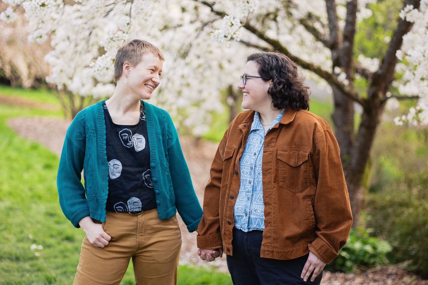 Two women holding hands and laughing together beneath blooming white cherry blossom trees in spring.