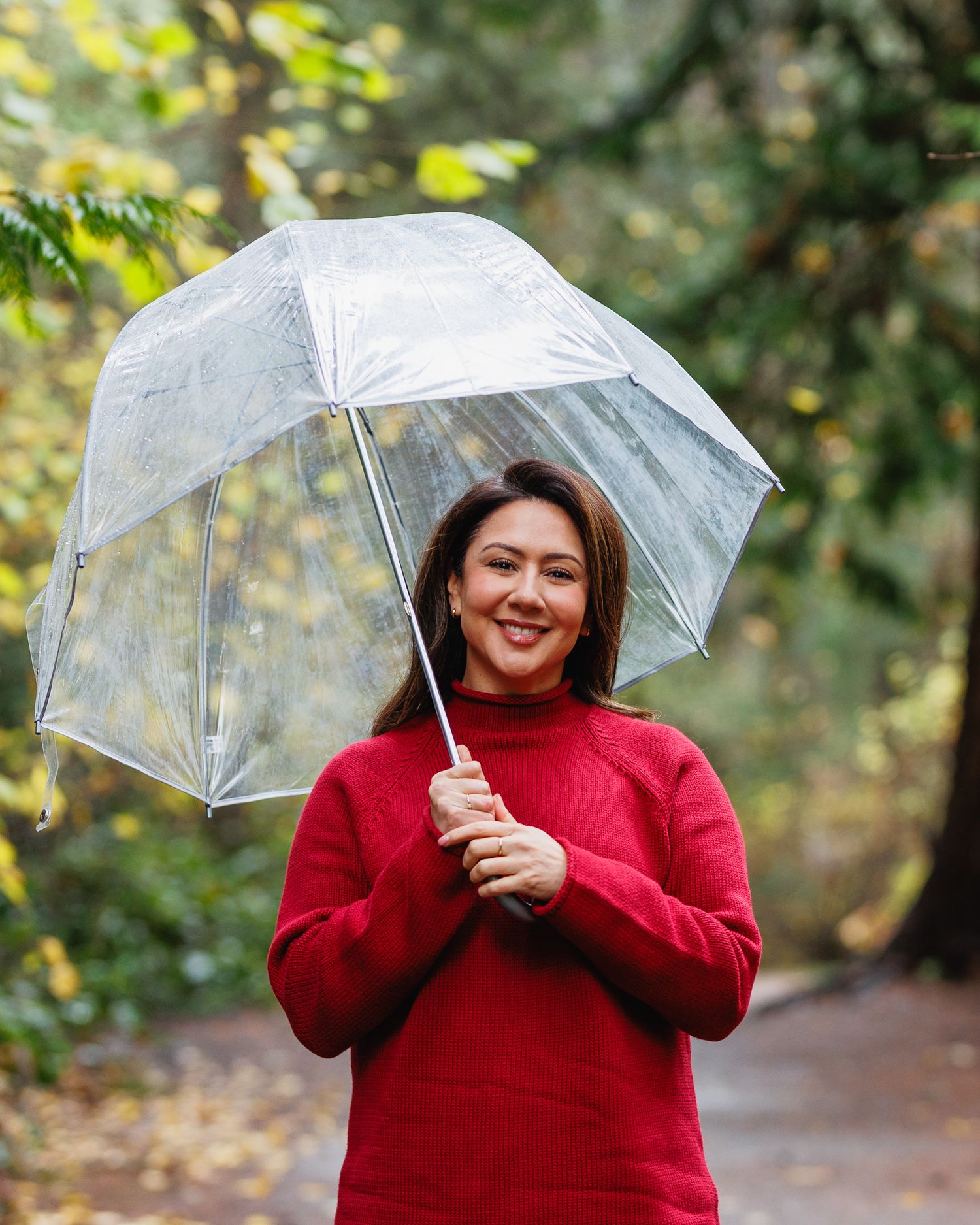 Smiling woman in red turtleneck sweater holding clear umbrella on tree-lined path in autumn park.