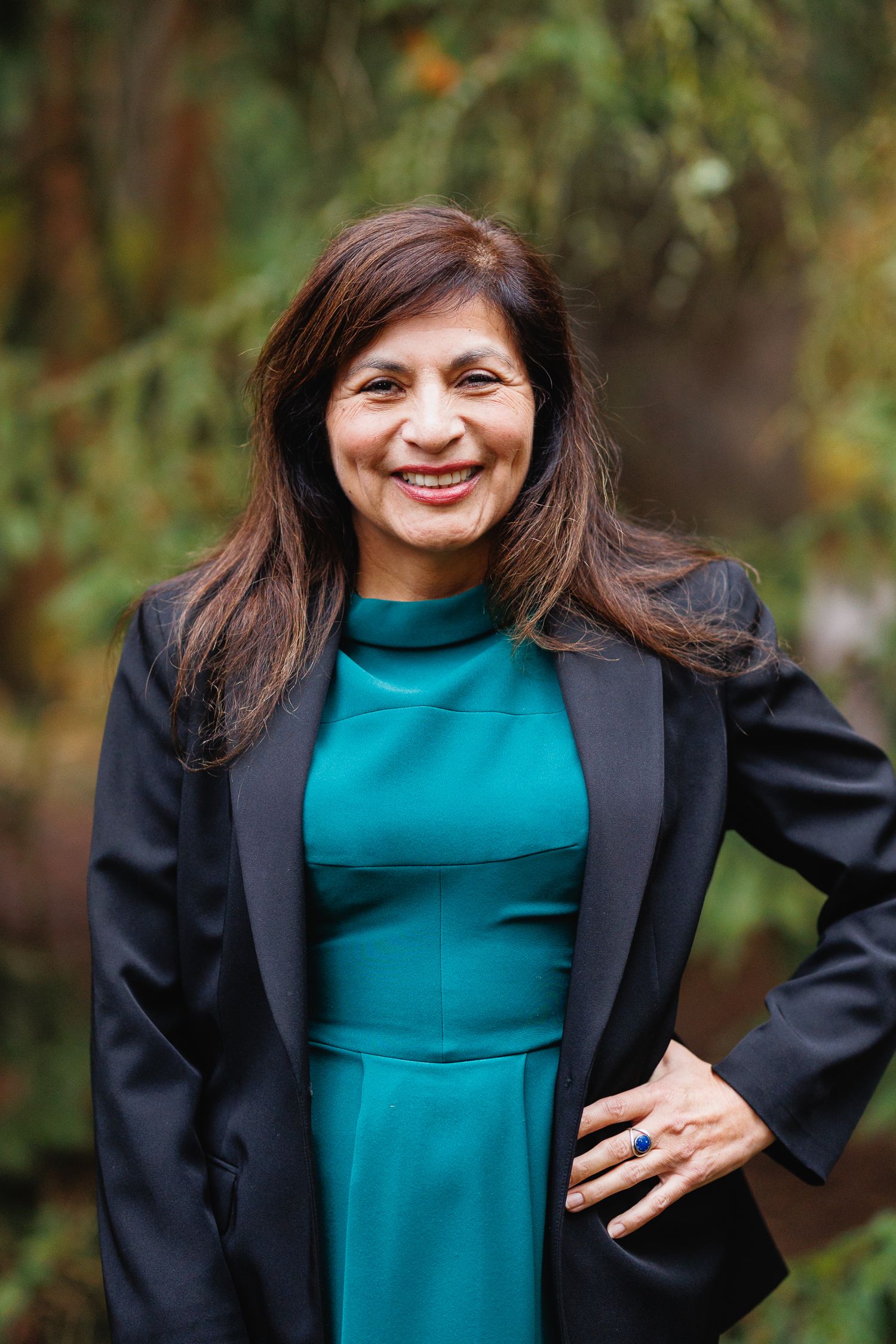 Confident woman in teal dress and black blazer smiling outdoors with green foliage background.