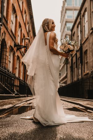 A bride in an elegant button-back wedding gown and flowing veil holds a peach bouquet on a city street.
