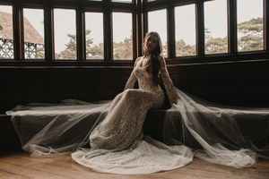 A bride in a flowing white wedding gown poses near large windows in a rustic industrial venue.