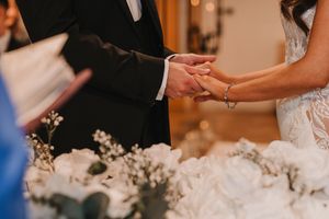 Close-up of bride and groom holding hands during wedding ceremony with white floral decorations in background.
