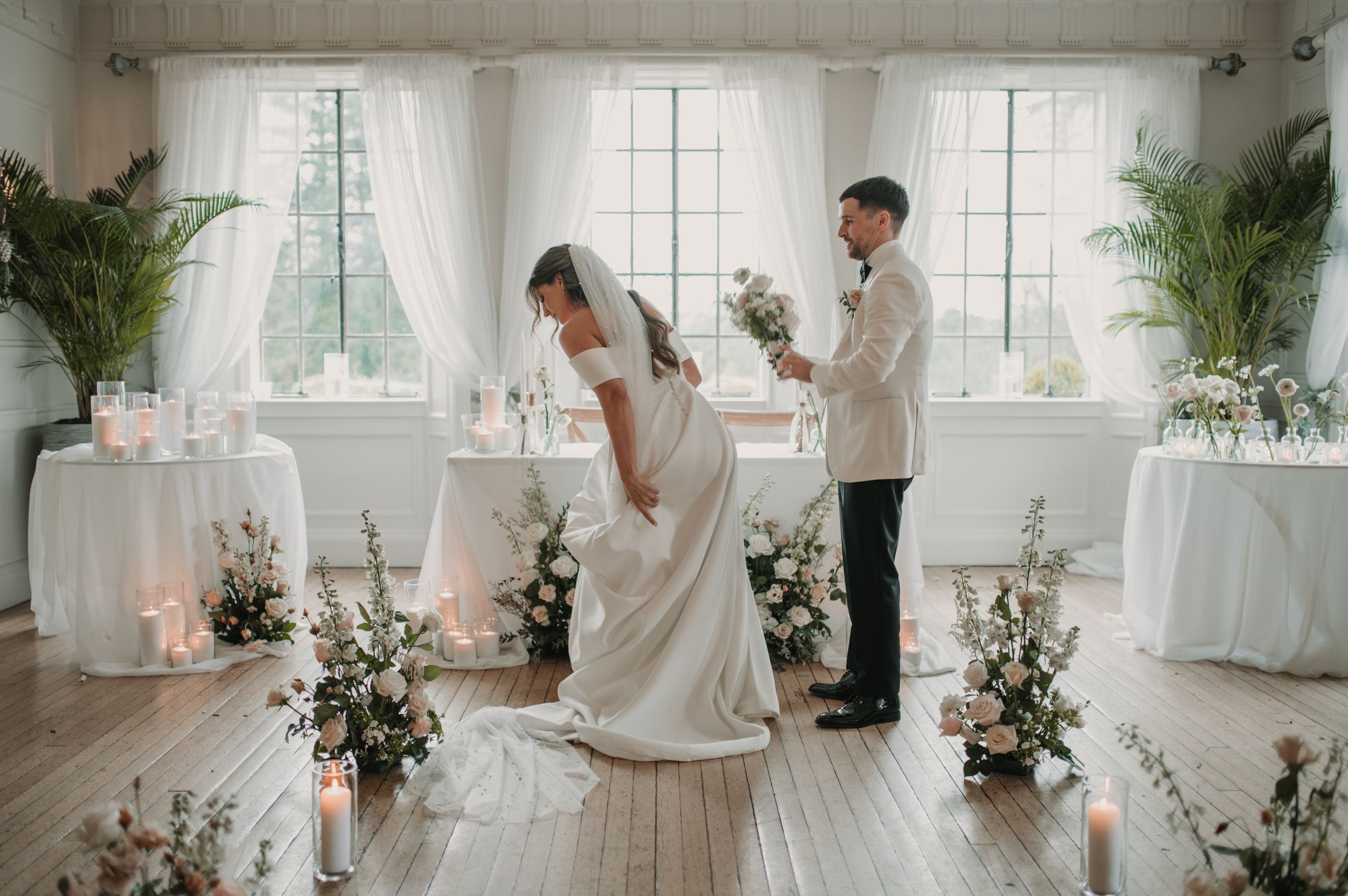 A bride and groom share an intimate moment in a bright white venue decorated with palm plants and floral arrangements.