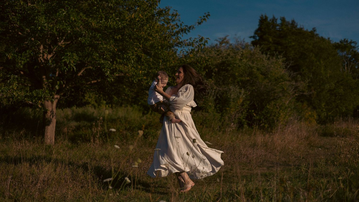A couple sits together on a blanket in a grassy field during golden hour sunset.